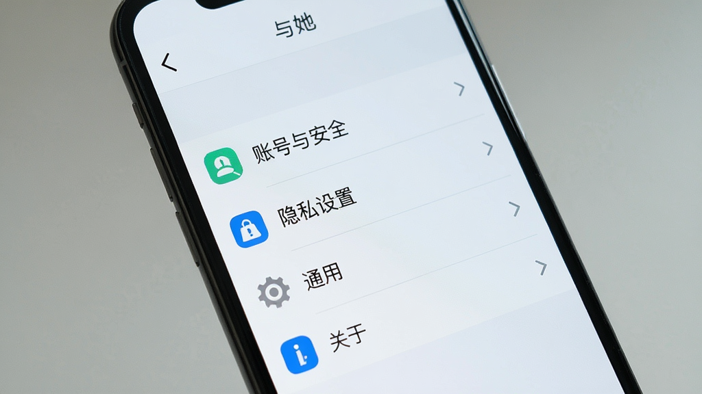 手机屏幕显示「与她」应用的设置菜单界面,菜单项包括「账号与安全」、「隐私设置」、「通用」、「关于」,界面简洁,布局为列表式,风格为清爽浅色系,细节包括菜单图标和分隔线,画质清晰干净,无多余元素
