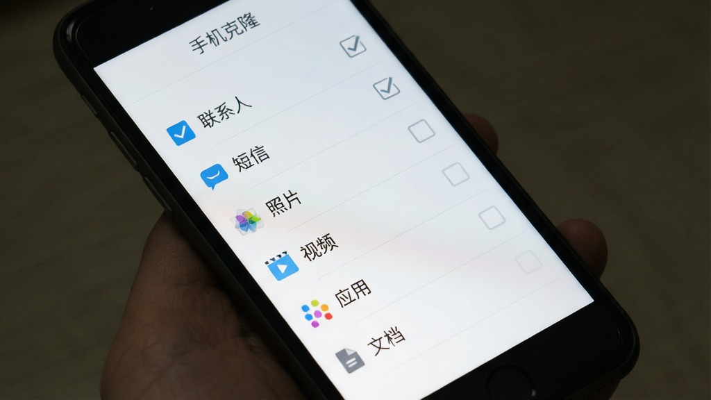 手机屏幕中显示一个勾选列表,列表项包括“联系人”“短信”“照片”“视频”“应用”“文档”等,每个项目前有勾选框,部分已勾选,屏幕上方有“手机克隆”标题,背景为干净的白色到浅灰色渐变,扁平化UI风格,细节清晰,画质干净,无多余元素