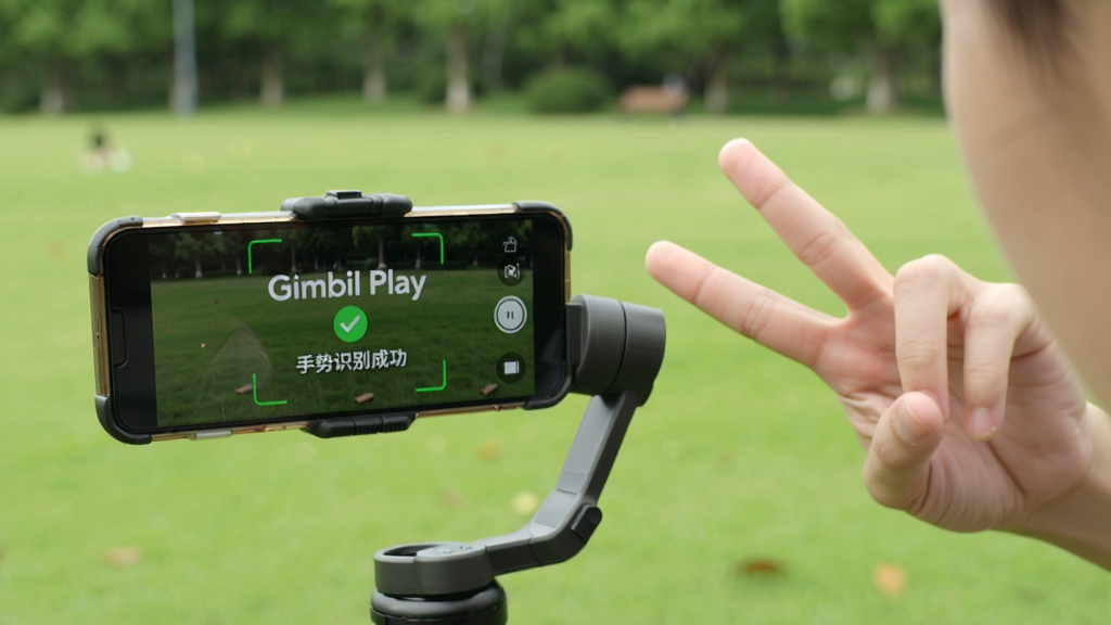 一个人对着手机云台比出剪刀手,屏幕显示 Gimbal Play 手势识别成功提示,背景为公园草地,主体为人物与云台,布局人物偏右,排版自然,风格生活写实风,细节包括手势动作、屏幕识别框、绿色对勾标识,画质高清,干净无多余元素