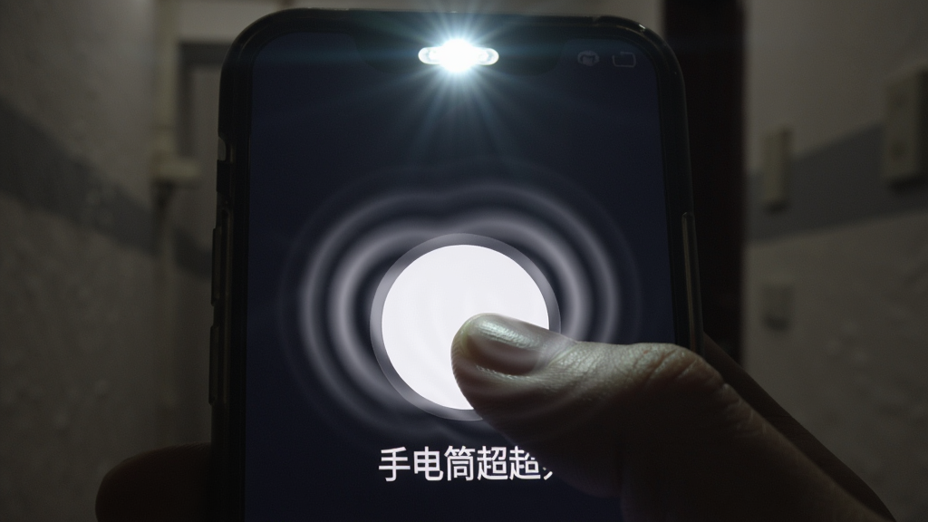 画面主体是一只手正握着手机,大拇指刚刚按下屏幕中央一个发着白光的巨大圆形按钮,按钮下方写着“手电筒超超亮”字样。布局采用特写镜头,手和手机占据画面大部分空间。排版上,按钮位于屏幕视觉中心,光线从手机顶部闪光灯射出。风格是真实生活场景结合科技光效,背景为昏暗的走廊或房间。细节上,手指按压按钮的瞬间有轻微的光晕涟漪扩散,闪光灯射出的光线照亮了前方的一小片区域,可以看到墙壁的纹理。画质清晰,画面干净,无多余元素。