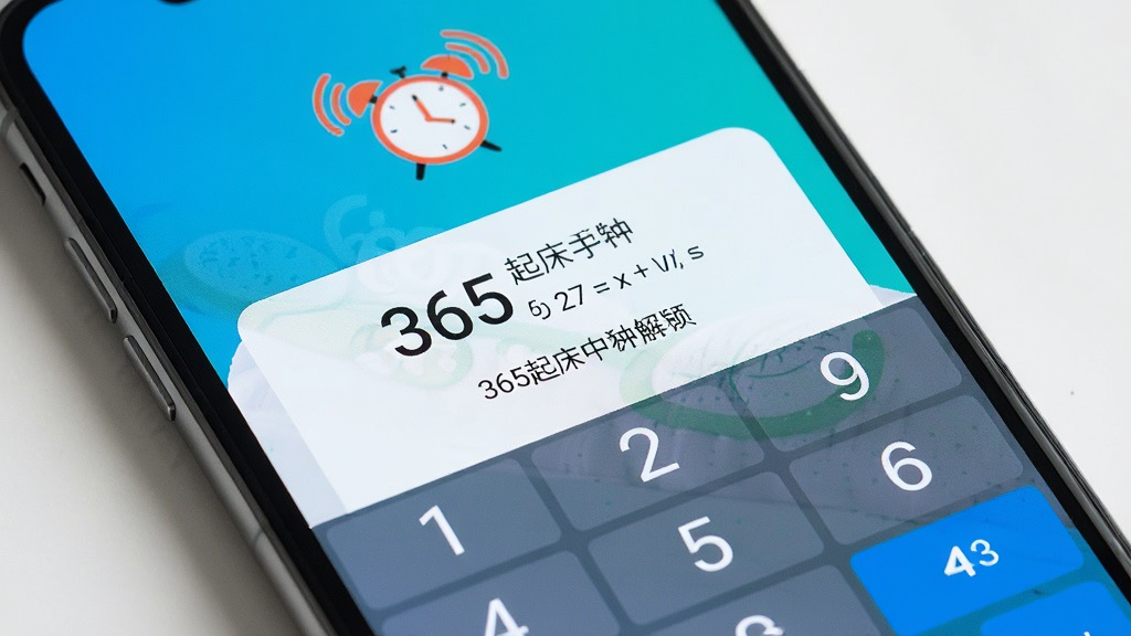一张手机屏幕特写图,画面中365起床闹钟弹出数学题解锁界面,上方显示闹钟正在响铃的图标,下方有数字键盘和题目,界面风格清爽,配色明快,画质清晰,干净,无多余元素
